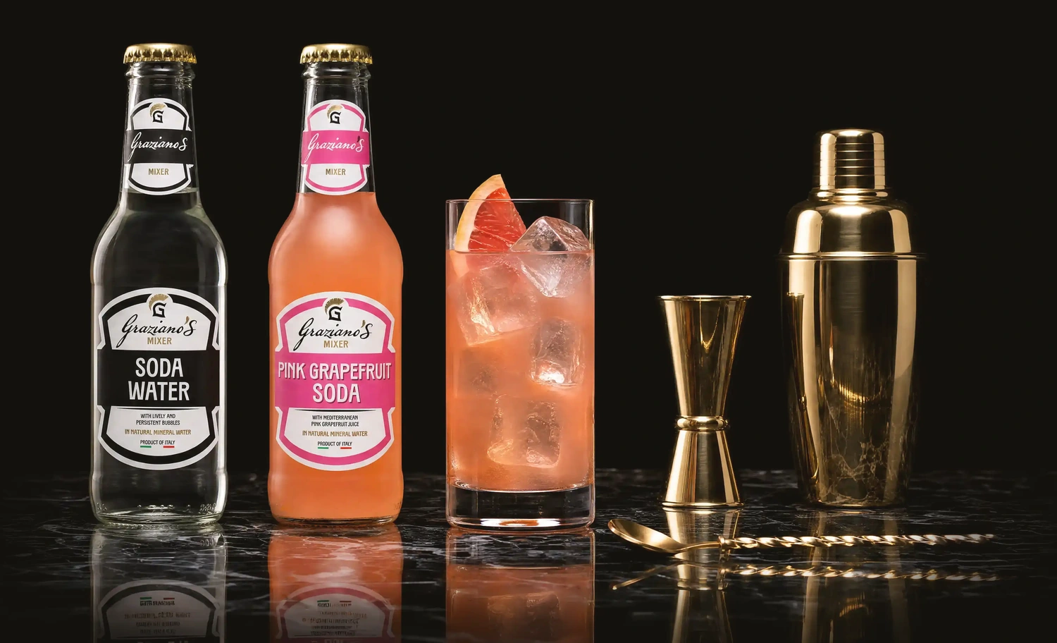 Bottiglie Soda Water e Pink Grapefruit Soda Graziano's con cocktail al pompelmo rosa, jigger e shaker dorati sul banco di un cocktail bar